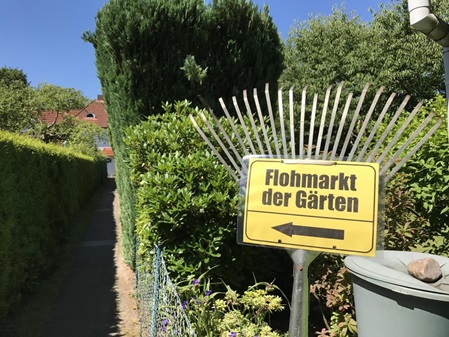 Ein Hauszuweg von der Straße zum Reihenhaus fotografiert. Der Weg ist um die 60m lang, rechts am Weganfang steht eine alte Harke/Rechen mit einem gelben DIN A4 Shilt wo in schwarz draufsteht: Flohamrkt der Gärten und Pfeil zum Haus. Vom Haus im Hintergrund sieht man nur etwas rotes Dach sowie viel Hecke den Weg runter und paar Büsche und Bäume.