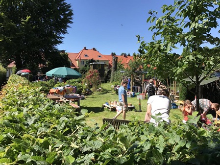 Vom Wirtschaftsweg hinter den Gärten über dessen Hecke in einen Garten fotografiert wo links und rechts im Garten Flohmarktstände zu sehen sind ebenso ein paar Besucher und im Hintergrund eine Reihenhausreihe der Siedlung. Der Himmel ist hellblau.