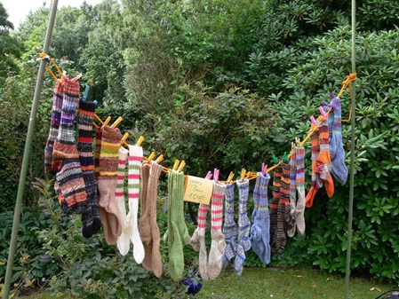 eine kurze Wäscheleinen an zwei grünen Wäschestangen behangen mit diversen bunten Socken am Wiesenende vor Büschen und Bäumen fotografiert.