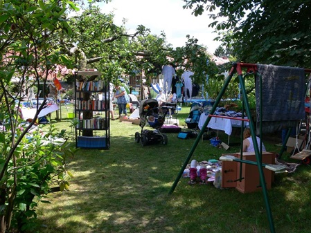 In den Garten fotografiert wo man diverse Flohmarktartikel auf Tischen und Regalen mitten von Bäumen und Büschen sehen kann ebenso ein paar wenige Personen und einen Kinderwagen.