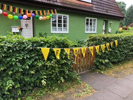 Ein grünes Siedlungs Doppelhaus schräg von link snach rechts zum Eingang fotografiert. Es hänen verschieden bunte Gierlanden über der Haustür und an der Gartenpforte auf der steht Flohmarkt.