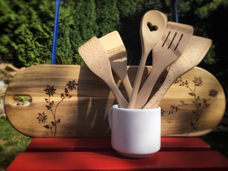 Holzkochlöffel und Pfannenwender stehen in einem weißen Keramikbecher auf einem roten Tisch im Garten vor einer Hecke. Hinter dem Becher steht ein Holzbrenn mit Blumen-Brandmuster.
